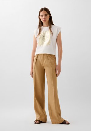 Top bianco senza maniche con stampa grafica oro; pantaloni a gamba larga beige; tessuto morbido; vestibilità casual con trame sottili.