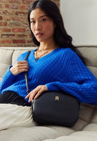 Borsa a tracolla nera in pelle con hardware dorato, texture liscia e dettagli con logo, abbinata a un maglione azzurro a trecce e una gonna plissettata.