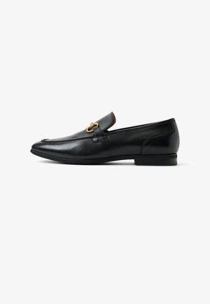 Mocasín de cuero negro con detalle de broche metálico dorado y tacón bajo, mostrado en perfil lateral sobre fondo blanco.