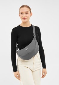 Borsa a tracolla grigia realizzata in tessuto morbido, di forma curvata, con tracolla regolabile e una patch con logo, indossata a spalla con una maglietta nera semplice.