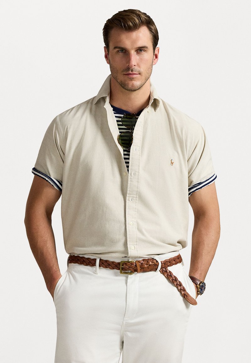 Beige short-sleeve button-up shirt with navy striped accents, worn over a navy and white striped T-shirt, paired with white pants and a braided brown belt.