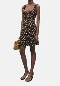 Musta kukkamekko ohuilla olkaimilla, tiukka bodice, leveähelmainen hame ja vaaleanpunaisia sekä keltaisia kukkakuvioita. Malli pitää käsissään kudottua vaaleanbeigeä laukkua.