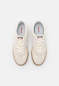 Zapatillas Levi's con parte superior en crema, acentos en cuero blanco y detalles en gamuza beige. Presentan una suela de goma y cordones beige. Logo rojo en la lengüeta.