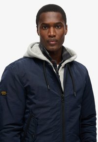 Marineblauwe bomberjack met een grijze capuchon, ritssluiting aan de voorkant en zwarte trekkoorden. Voorzien van een patch met "Superdry" op de linkermouw.