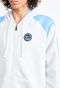 Sweatshirt branco com fecho éclair e detalhes em malha azul clara nos ombros. Apresenta um logótipo da Nike em hexágono preto no peito. Material em mistura de algodão.