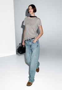 T-shirt in cotone grigio con bordi bianchi, jeans in denim blu loose, scarpe in suede marroni e una piccola borsa nera, posati su uno sfondo neutro.