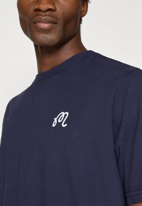 Zilā kokvilnas T-kreklā ar baltu izšūtu burtu "m" uz krūšu, apaļa kakla un īsām piedurknēm.