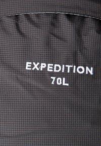 Tmavě šedá látka s texturovaným vzorem, obsahující bílý vyšívaný nápis "EXPEDITION 70L." Design klade důraz na užitečnost a kapacitu úložného prostoru.