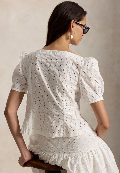 Γυναίκα με σκούρα μαλλιά που φορά γυαλιά ηλίου πλούσιου χρώματος, λευκό τοπ με φουσκωτά μανίκια και ταιριαστή φούστα, καθισμένη σε ξύλινο σκαμνί με την πλάτη προς τα εμπρός.