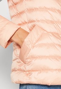 Jasnoróżowa puchowa kurtka z pikowanym wzorem, zbliżenie na rękaw i detal guzika przy mankiecie. Gładki, błyszczący materiał.