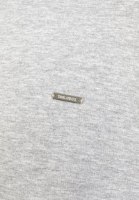 Sweat-shirt gris avec une texture douce comportant une étiquette en métal portant l'inscription « SHILODOX » près du centre, présentant un design minimaliste.