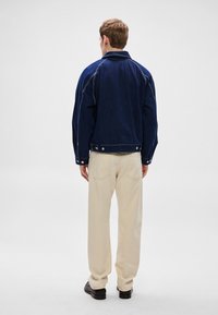 Marineblå denimjakke med tettsittende design, kontrastsømmer og trykknapper; kombinert med kremfargede bukser og svarte sko.