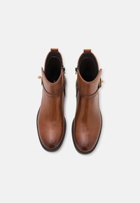 Bottes de cheville en cuir marron avec une finition brillante, fermeture éclair sur le côté et une boucle dorée décorative. Semelle noire plate, design épuré.