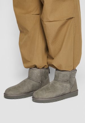 Bottines en daim gris portées avec un pantalon ample beige sur un fond clair uni.
