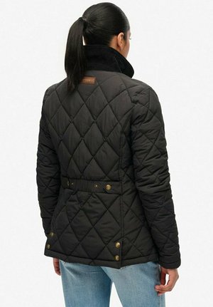 Vrouw met een zwarte paardenstaart, gekleed in een zwarte gewatteerde jas met knoopdetails en lichtblauwe jeans, gezien van achteren.