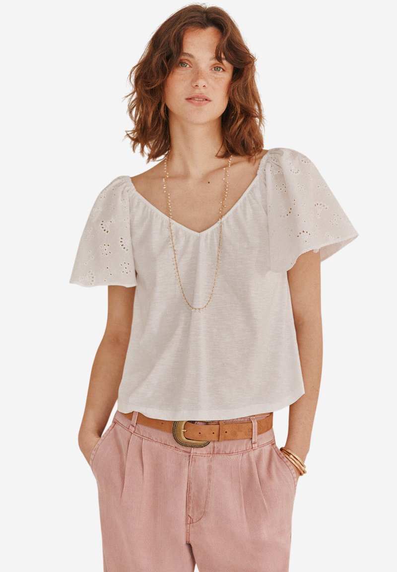 Top bianco in cotone con maniche a sbuffo e dettagli ad occhiello, scollatura a V e vestibilità comoda. Indossato con pantaloni rosa chiaro e una cintura marrone.