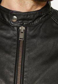 Černá kožená bunda s vysokým límcem, vybavená kovovým zipem a patentním knoflíkem. Hladká textura s viditelnými švy.