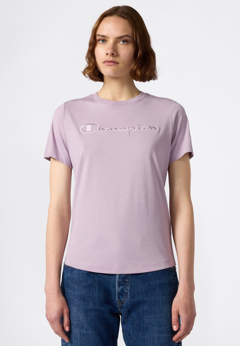 Jasnofioletowy T-shirt z krótkim rękawem, z haftowanym logo "Champion" w ciemniejszym odcieniu fioletu. Gładki materiał bawełniany z okrągłym dekoltem.