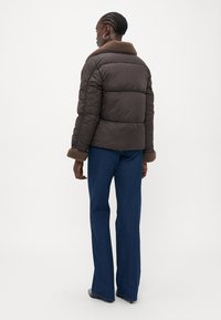 Brun puffer-jakke med en beige pelskrage, ribbede mansjetter og en flat silhuett. Kombinert med vide blå jeans og sorte sko.