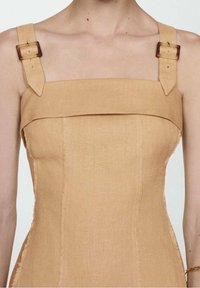 Robe en lin beige avec larges bretelles dotées de fermetures à boucle réglables. Texture lisse avec coutures visibles et corsage structuré.