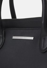 Gros plan d'un sac à main en cuir noir avec deux anses et une petite plaque de logo Calvin Klein en argent sur le devant.