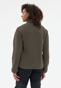 Brown fleece jacket with a high collar and flat knit shoulder panel. Features textured fabric and fitted sleeves. Worn with black pants.