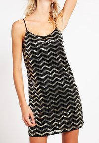 Kobieta w bezrękawnej czarnej mini sukience z błyszczącym srebrnym wzorem w zygzaki i cienkimi ramiączkami.