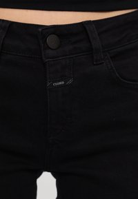 Zone de la taille d'un jean en denim noir avec une fermeture à bouton et une petite étiquette portant l'inscription "CLOSED" près de la fermeture éclair.
