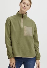 Maglione in pile verde oliva con mezza zip frontale, tasca con zip beige e polsini elasticizzati; texture morbida, vestibilità comoda.