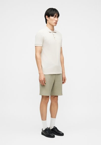 Νεαρός άνδρας που στέκεται, φοράει μπεζ πόλο πουκάμισο, ανοιχτό καφέ σορτς, λευκές κάλτσες και μαύρα αθλητικά παπούτσια μπροστά σε απλό λευκό φόντο.