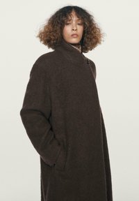 Žena s kudrnatými vlasy, oblečená v tmavě hnědém oversize vlněném kabátu s vysokým límcem, stojící na neutrálním světle pozadí.