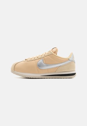 Beige Nike-sneakers med silverfärgad swoosh-logga, snörningsdesign och räfflad sula visad från sidan på vit bakgrund.