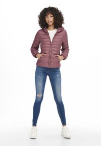 Doudoune violette rembourrée avec zip sur le devant, capuche et poches. Portée par-dessus une chemise blanche et un jean skinny bleu déchiré, associée à des baskets blanches.