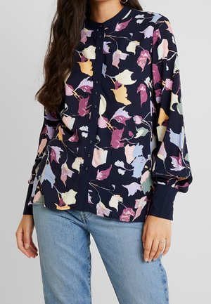 Woman wearing a navy button-up blouse with pastel floral leaf print and light blue jeans, standing against a plain light background.