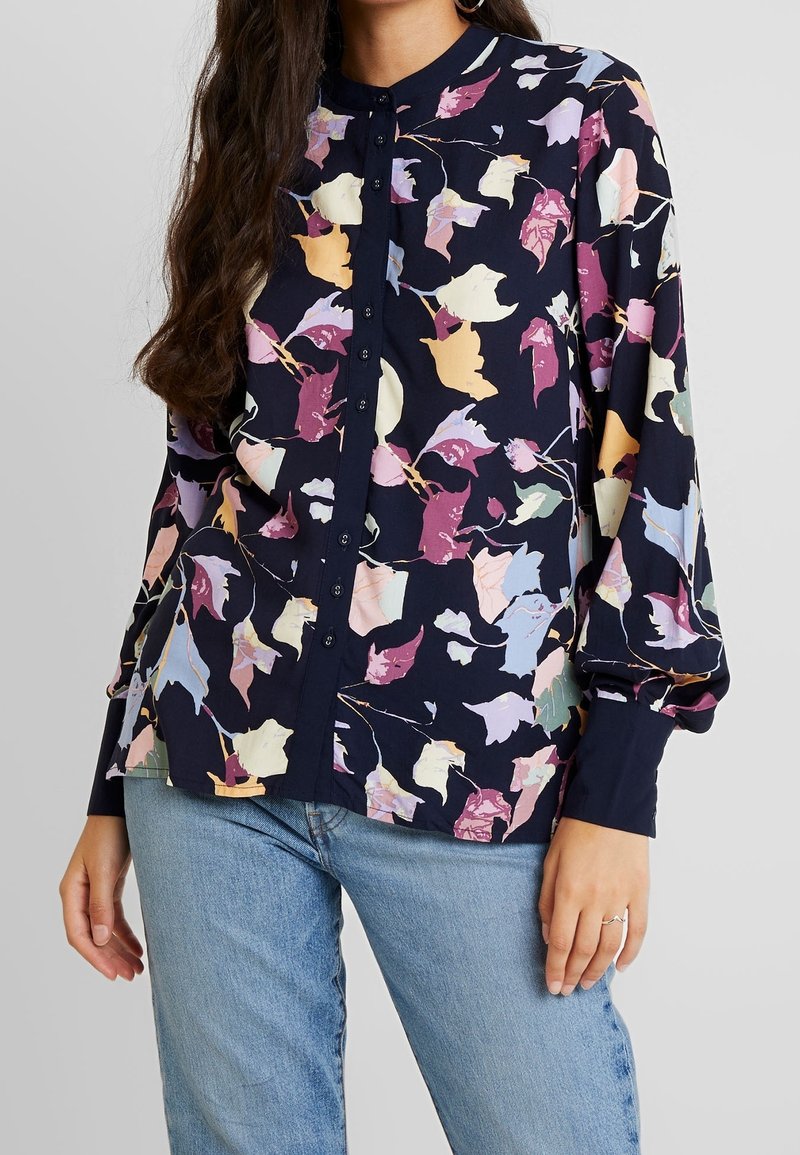 Femme portant un chemisier bleu marine à boutons avec un imprimé floral pastel et des feuilles, et un jean bleu clair, debout devant un fond uni clair.