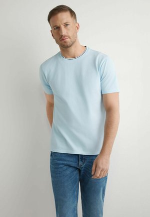 Mężczyzna stojący na białym tle, ubrany w jasnoniebieski t-shirt z krótkim rękawem i niebieskie dżinsy, patrzący prosto przed siebie.