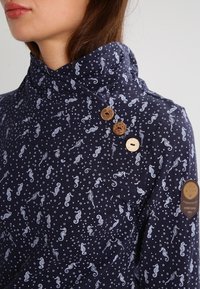 Marinblus med vitt sjöhästs- och bubbelmönster, hög krage med tre träknappar, brun rund lapp på överarmen, påburen av en person.