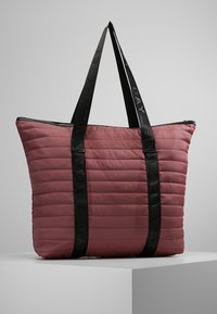 Gevoerde roze draagtas met zwarte logo handvatten. Heeft een gequilt ontwerp, ritssluiting en rechthoekige vorm. Gladde textuur.