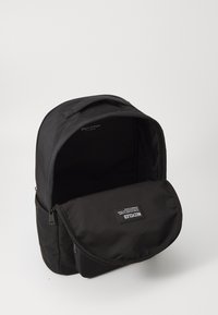 Zwarte rugzak van duurzaam materiaal, met een rits hoofdcompartiment en een klep aan de voorkant met een label, minimalistisch ontwerp.
