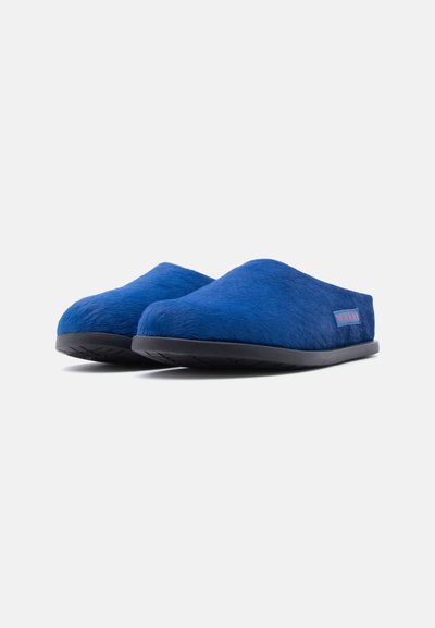 Chaussons bleu doublés de fourrure avec une texture douce, une forme arrondie et une semelle en caoutchouc noir. Ils comportent une petite étiquette rouge et bleue sur le côté.