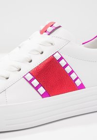 Baskets blanches avec une bande texturée rouge et rose, ornées de clous métalliques et de lacets plats blancs. Semelle épaisse blanche pour plus de hauteur.
