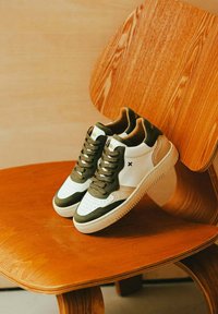 Baskets blanches et vert olive avec un bout perforé, des accents en cuir texturé et des lacets ronds reposant sur une chaise en bois.