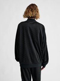 Persona con capelli mossi fino alle spalle che indossa una giacca nera larga a maniche lunghe e pantaloni abbinati, in piedi di spalle su uno sfondo semplice.