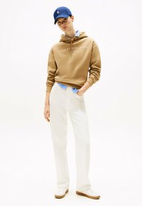 Sweat à capuche en coton marron avec logo blanc, chemise à rayures bleues en dessous, pantalon blanc et baskets blanches avec semelles marron clair, le tout couronné d'une casquette bleue.