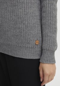 Grauer gestrickter Pullover mit strukturiertem Muster, gerippten Bündchen und einem Holzlogo-Tag am unteren Saum. Trägt man über schwarzer Kleidung.