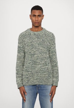 Groene en crèmekleurige gebreide trui met een ronde hals, textuurpatroon en lange mouwen. Gecombineerd met lichtblauwe denim jeans.