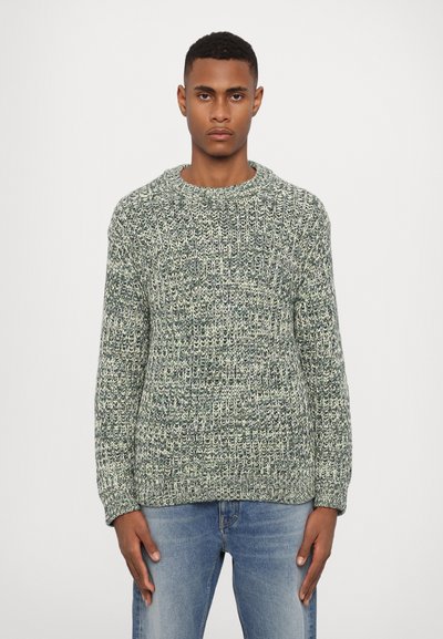 Grüner und cremefarbener Strickpullover mit Rundhalsausschnitt, strukturiertem Muster und langen Ärmeln. Kombiniert mit hellblauen Jeans.