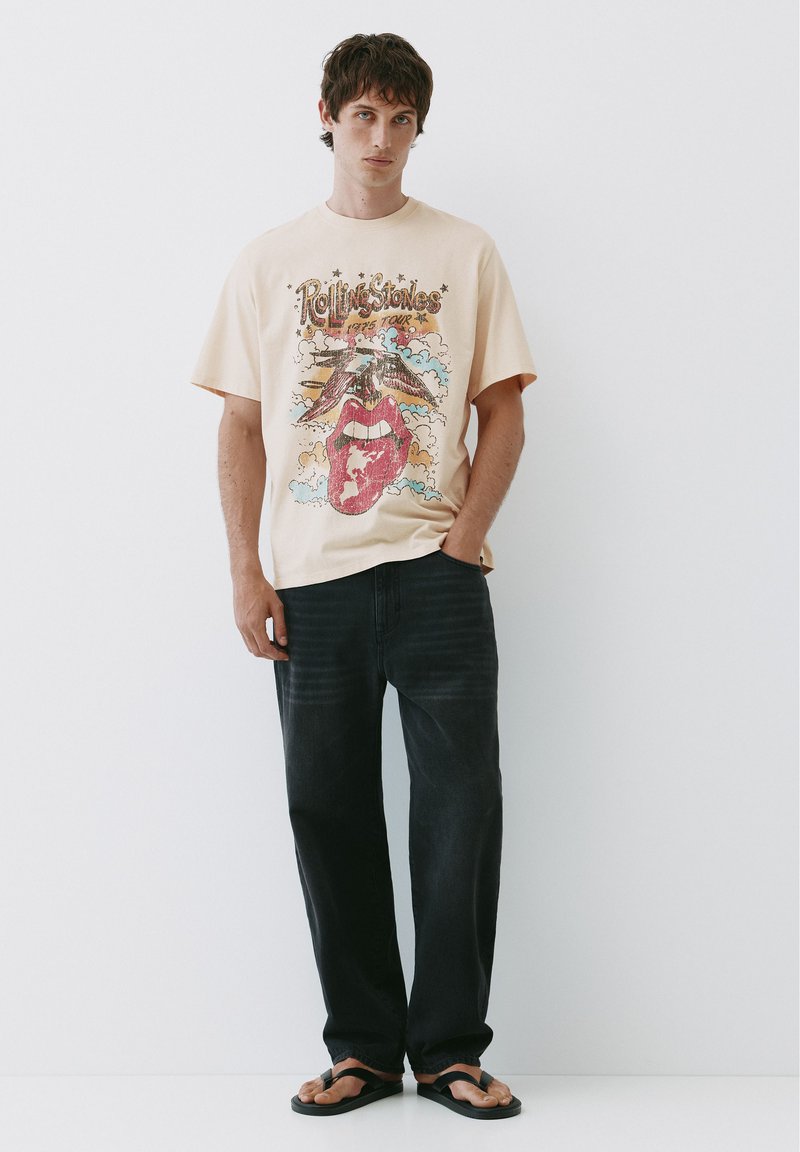 Jeune homme aux cheveux courts et foncés portant un t-shirt beige à motif Rolling Stones, un jean noir et des tongs noires, debout contre un mur blanc.