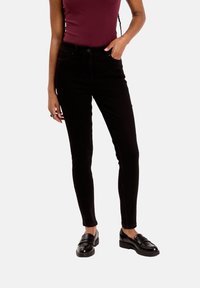 Jeans noirs slim avec des mocassins noirs et un top sans manches bordeaux, avec une main dans la poche, sur fond blanc.
