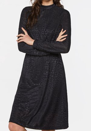 Femme portant une robe noire à manches longues avec une texture florale subtile, se tenant les bras croisés devant un fond uni.